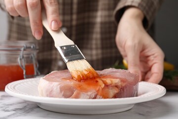 Woman spreading marinade onto raw meat with basting brush at white marble table, closeup