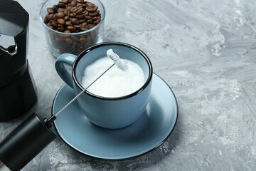 Mini mixer (milk frother), whipped milk in cup and coffee beans on grey textured table, closeup....
