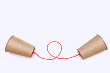 Two paper cups connected by orange thread isolated on white background close-up with free space for text. Cardboard kraft cup tied for toy phone line. Communication concept