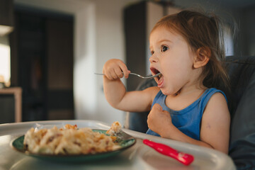Cute baby eating first solid food, infant sitting in high chair. Child tasting, discovering new food. Cozy kitchen interior. Healthy food concept.