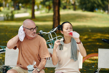 Mature couple sitting on park bench, resting after bicycle ride and drinking water