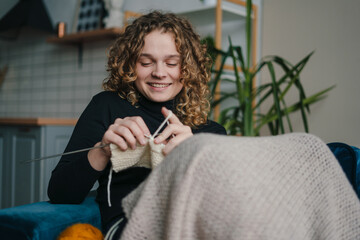 A young curly haired woman knitting a scarf in a cozy room on sofa. A cold fall or winter weekend...
