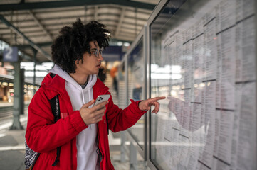 Curly-haired young traveler scrutinizing trains timetable and looking involved