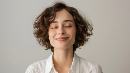 Portrait of a shy female corporate intern smiling with eyes shut against grey background