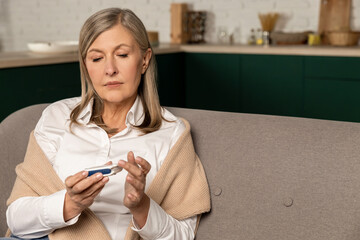 Good-looking mid aged woman sitting on the sofa and making blood sugar test