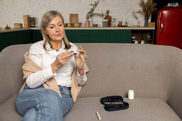 Mature woman sitting on the sofa and checking blood sugar with glucometer