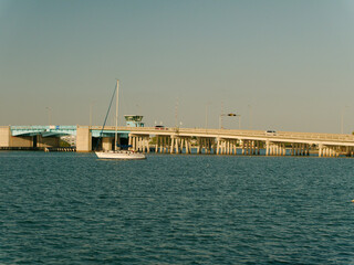 Fototapeta premium Wide View South East from St Pete Beach Community Center towards Corey Causeway drawbridge Boca Ciega Bay with sailboat no mast in blue water near sunset. In Florida. 