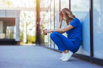Tired nurse sits on floor outdoors of the medicine clinic after a hard dutty. Exhausted tired doctor or nurse. Clinic and hospital medical stuff working over hours. Overworked professional. 