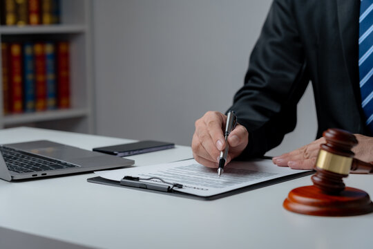 A lawyer in a suit is sitting at his desk, signing legal documents with a gavel on the side.