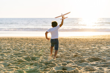 A joyful boy lifts a toy plane up high on the beach at dusk.