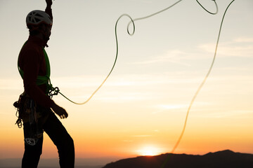 A man is standing on a mountain with a rope in his hand. The sky is orange and the sun is setting