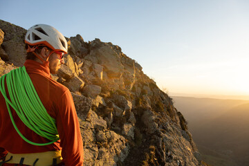 A man in an orange jacket is standing on a rocky mountain, looking out at the horizon. The sun is setting, casting a warm glow over the landscape. The man is wearing a helmet and a green rope