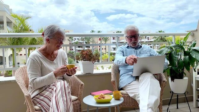 Candid capture of relaxed couple of seniors enjoying lifestyle in retirement, elderly 70 years old people sitting outdoors on terrace, reading a book, surfing the net, using phone, enjoying a snack