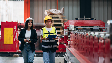 Two women wearing safety gear and holding clipboards walk through a factory