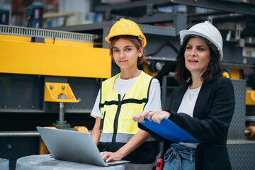 Two women wearing hard hats and safety vests stand next to a laptop computer