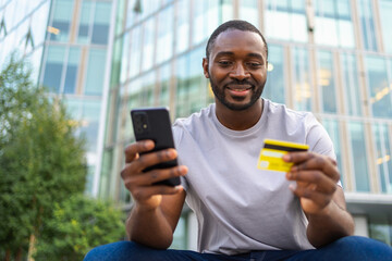 African American man shopping online holding smartphone paying with gold credit card Guy on urban...
