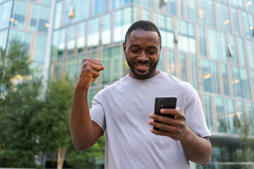 Happy African American man euphoric winner with smartphone on street in city. Person guy looking at cell phone reading great news getting good result winning online bid feeling amazed. Winning gesture