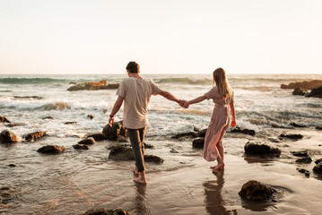 Husband and wife walking along the beach while holding hands and ocean waves crash along rocky shore