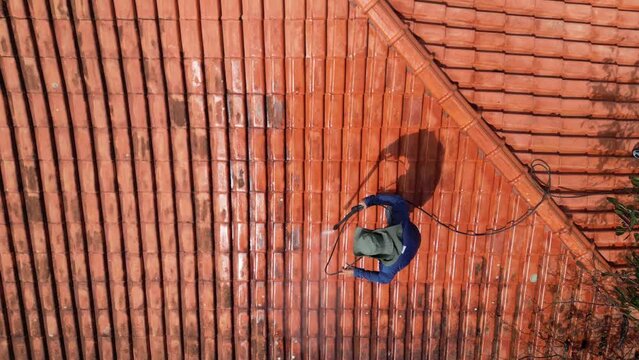 A person outdoors cleaning the red clay roof using a high pressure washer. Professional washing house tiles with water hose.