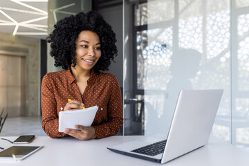 A young professional woman is engaged in a business meeting, taking notes in a modern office setting with natural light and a laptop in front of her.