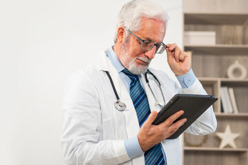 Mature senior male doctor in medical uniform using digital tablet in clinic. Therapist professional healthcare expert searching information online in hospital room. Medicine healthcare medical checkup