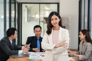 Confident young businesswoman with arms crossed standing in office with colleagues discussing in...