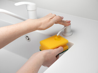 Female hand press on built-in dispenser in the sink with detergent. Woman washing dishes by yellow sponge with soap in white kitchen
