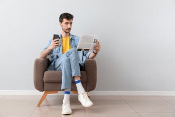 Handsome young man with mobile phone reading newspaper in armchair near light wall