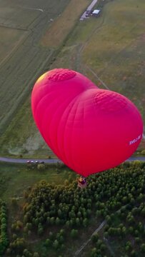 Red aerostat over fields. Romantic hot air balloon in a form of a heart travels in the sky on beautiful nature background at sunset. Top aerial view. Vertical video
