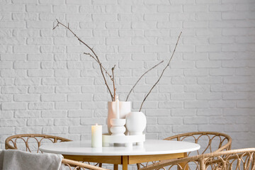 Stylish table with burning candles, plant branches and rattan chairs near light brick wall