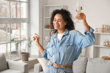 Young African-American woman with glass of champagne dancing at home