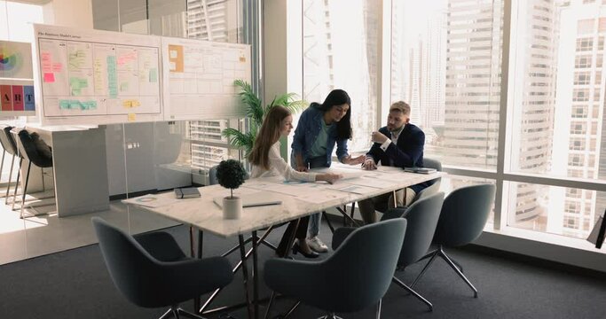 Three multi-ethnic colleagues, Indian and European teammates discussing infographic data and effectiveness of marketing strategy gathered in modern meeting room with city view. Market research, sales