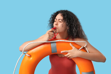 Beautiful young African-American female lifeguard with ring buoy whistling on blue background