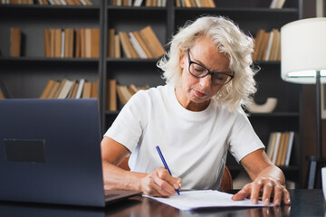 Middle aged senior woman using laptop computer writing notes at workplace. Focused mature old businesswoman work at office making written records doing online work. Mature boss professional worker
