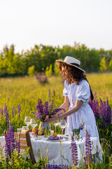 Beautiful happy young woman on a meadow arranging table for outdoor event, gathering wildflowers,...
