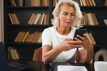 Confident stylish European middle aged senior woman using smartphone at workplace. Stylish older mature 60s lady businesswoman with cell phone in office. Boss leader using internet apps