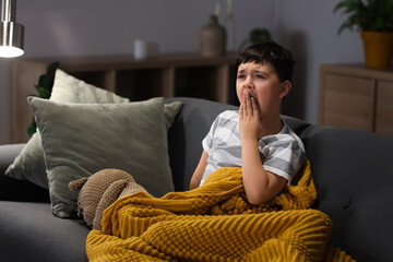 Cute sleepy little boy with teddy bear yawning on sofa in living room