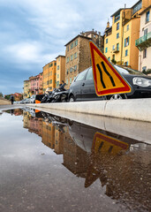 Road narrowing traffic sign, cars and houses in Menton reflect in a puddle after rain