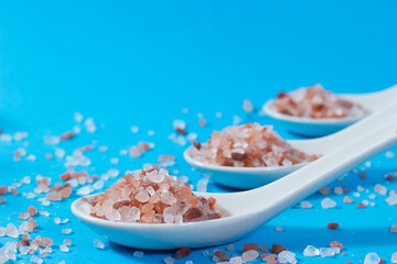 Close-up of ceramic teaspoons with pink Himalayan salt on blue background.