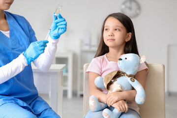 Little Asian girl with toy bear receiving vaccine in clinic