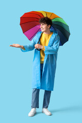 Young man in raincoat with rainbow umbrella on blue background