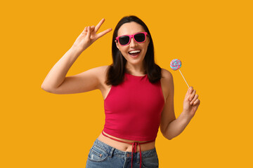 Fototapeta premium Beautiful young woman in sunglasses with sweet lollipop showing victory gesture on yellow background
