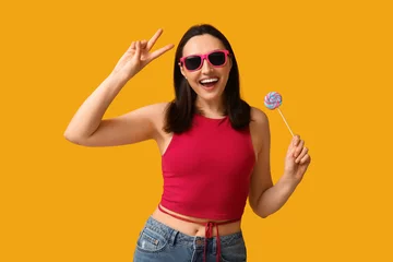 Foto op Aluminium Beautiful young woman in sunglasses with sweet lollipop showing victory gesture on yellow background © Pixel-Shot