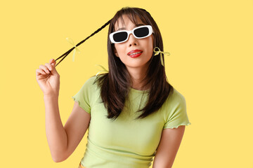 Stylish young Asian woman in sunglasses with bows on yellow background