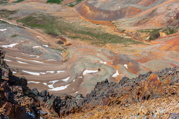 Scenic dizzy top-down view from precipice edge with sharp rocks to unusual multicolor stony valley with iron river in sunlight. Colorful awesome alpine scenery with snow in multi-color hilly valley.