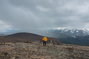 Man in yellow raincoat on stony pass with view to valley against large snow-capped mountain range in gray rainy low clouds. Guy among sharp stones. Big snowy mountains in rain under grey cloudy sky. - 786683923