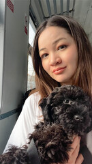 Cute Korean woman holding black poodle puppy sitting in train on railway trip. Traveling with dog. Asian female person in train car. Selfie with puppy. Soft focus. film grain pixel texture. Defocused.