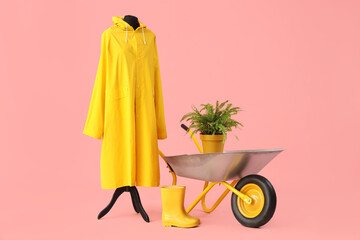 Raincoat, rubber boots, plant and wheelbarrow on pink background