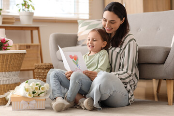 Happy mother with her cute little daughter and greeting card sitting on floor at home