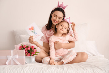 Obraz na płótnie Canvas Happy mother with greeting card and her cute little daughter in crowns sitting on bed at home